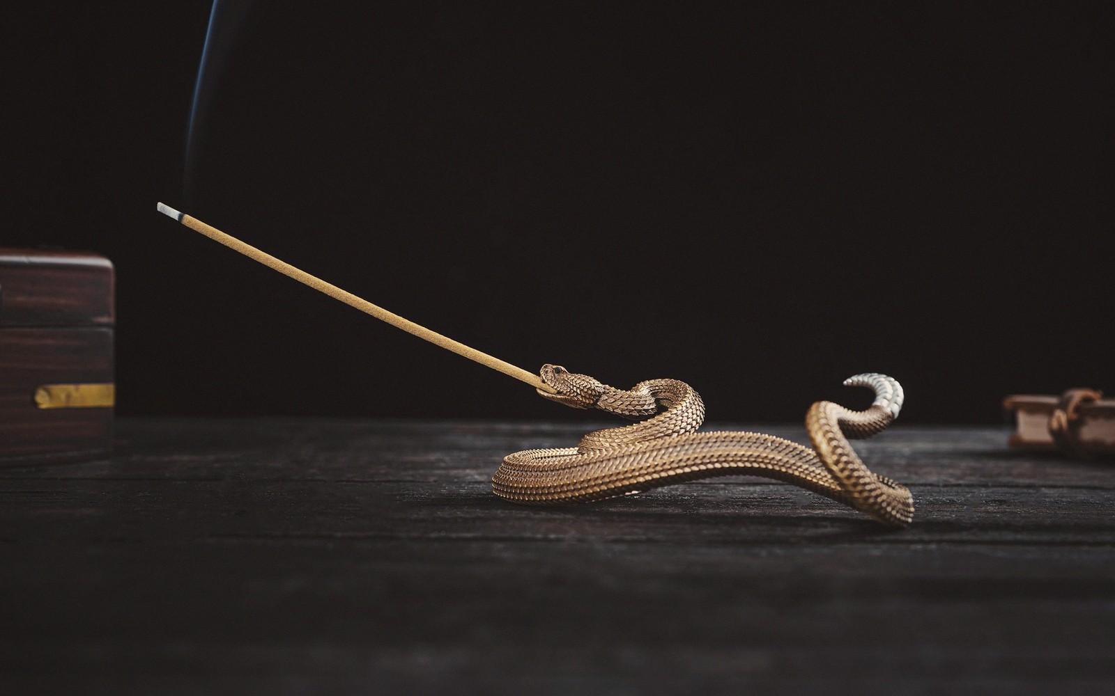 A coiled brass snake incense holder supports a smoking incense stick on a dark wooden table
